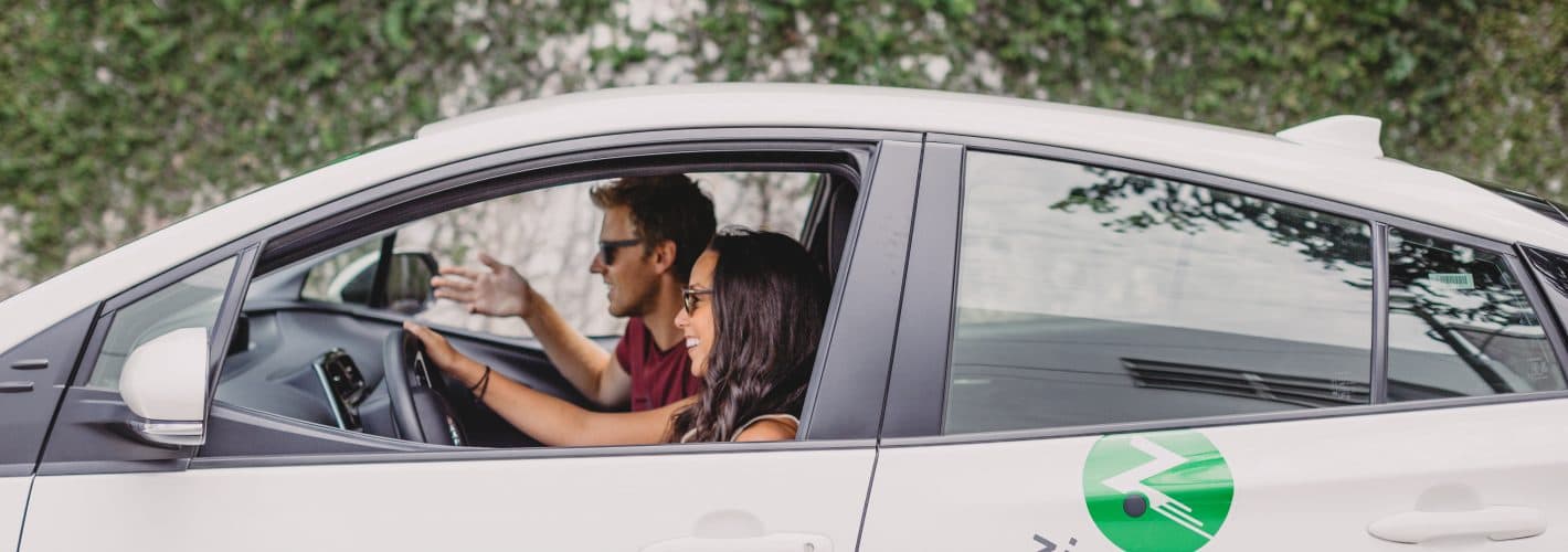 A woman driving a white Zipcar with a man in the passengers seat